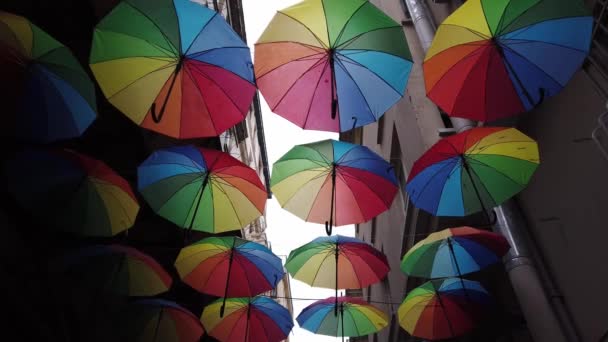 Muchos coloridos paraguas abiertos arco iris cuelgan sobre estrecha calle entre casas - Imágenes, Vídeo