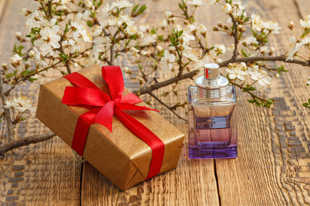 Подарочная коробка и бутылка духов с ветками цветущей вишни на деревянном фоне. Концепция дарения подарков в праздничные дни. - Фото, изображение