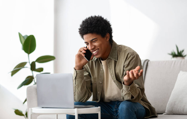 Концепция обучения. Положительный черный подросток говорит по мобильному телефону возле ноутбука дома - Фото, изображение