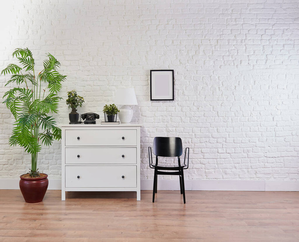 Tuğla duvar, lamba, çerçeve ve vazo önünde dekoratif beyaz dolap ve siyah sandalye. - Fotoğraf, Görsel