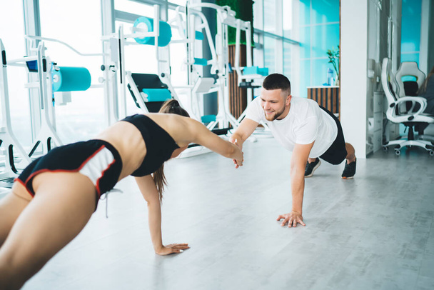 Cheerful powerful sportspeople in activewear standing in plank position and holding hands while having cardio workout in spacious light gym together - Fotoğraf, Görsel