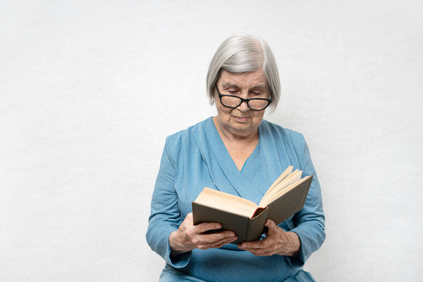 80-as évekbeli nő ősz hajjal és ráncos bőrrel olvas egy könyvet. Szemüveg van rajta..  - Fotó, kép
