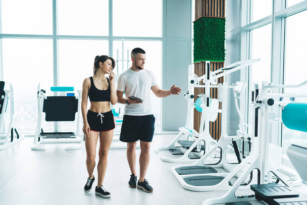 Bearded personal trainer with tablet in hand walking with young female in sportswear and explaining about gym equipment in modern fitness studio - Foto, Imagem