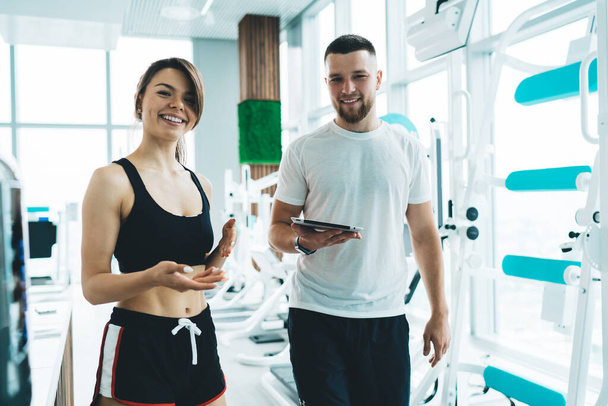 Smiling fit woman in black sportswear with bearded male personal trainer with tablet in hand standing in bright modern gym - Φωτογραφία, εικόνα