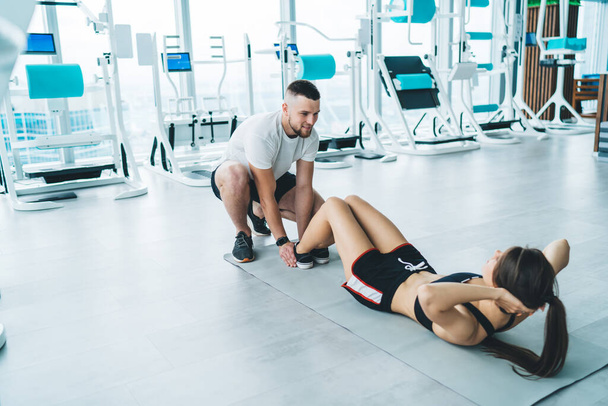 Full body of unrecognizable sportswoman in activewear doing abs exercise on fitness mat with personal trainer in modern light gym - Foto, Imagem