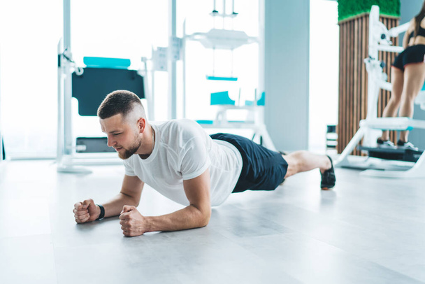 Full body of powerful bearded sportsman in activewear doing plank exercise on floor while having workout in modern light gym - Fotó, kép