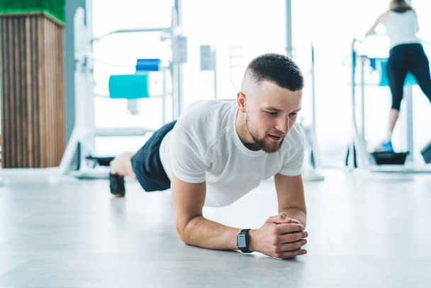Focused sportsman in smart watch doing elbow plank in middle of modern gym during intense warm up in bright daylight - Foto, Bild
