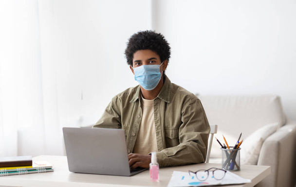 Remote education during COVID-19 outbreak. Black teen guy in protective mask using laptop computer for online schooling - Photo, Image