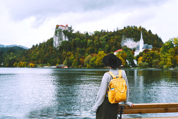 Beautiful woman looking at Lake Bled with Castle and Church in background. Tourist travel in Slovenia, Europe. Boho style hipster girl relaxing in nature outdoor near by lake on foggy and rainy day. - Zdjęcie, obraz