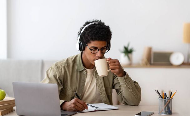 Afrikkalainen amerikkalainen teini kuulokkeissa opiskelee etänä kotoa, juo kahvia ja tekee muistiinpanoja copybookissa - Valokuva, kuva