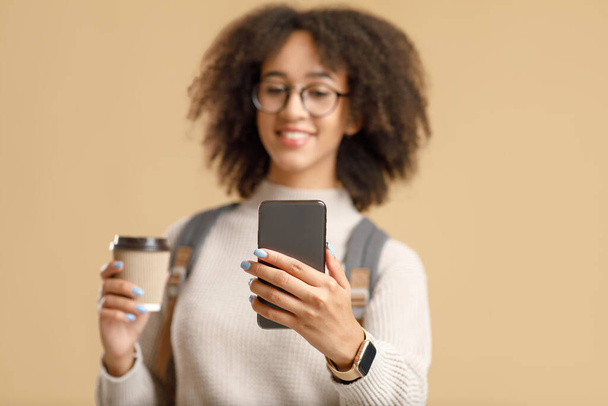 Fokus auf Smartphone mit Leerraum in den Händen einer afrikanisch-amerikanischen Frau mit Rucksack und Brille, die eine Tasse Kaffee hält - Foto, Bild