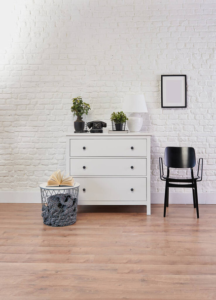 Decoratieve witte kast en zwarte stoel voor de bakstenen muur, lamp, frame en vaas van plantaardige decoratieve stijl. - Foto, afbeelding