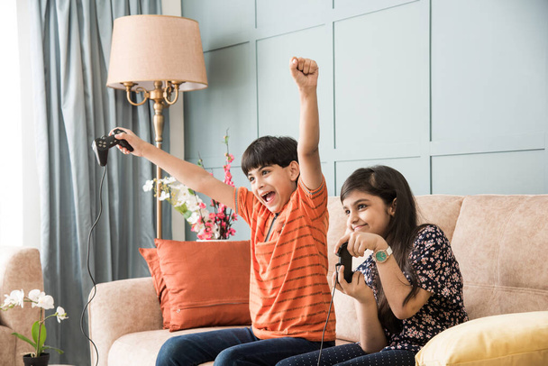 Cheerful Indian Asian kids playing video game using joystick or controller while sitting on sofa - Foto, afbeelding