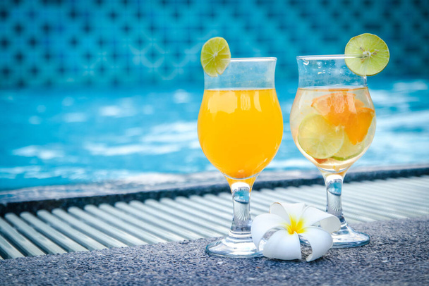 Δύο ποτήρια χυμό πορτοκαλιού, μαζί με λουλούδια Plumeria, τοποθετούνται στο χώρο της πισίνας τα Σαββατοκύριακα.. - Φωτογραφία, εικόνα