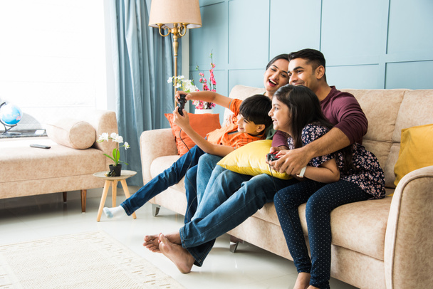 Индийская молодая семья из четырех человек играет в видеоигры с помощью контроллера или джойстика, сидя на диване - Фото, изображение