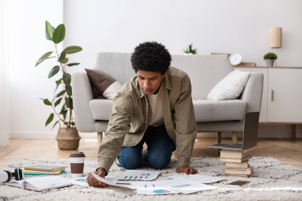 Full length portrait of black teen guy arranging materials for his coursework paper on floor at home - Foto, Imagem