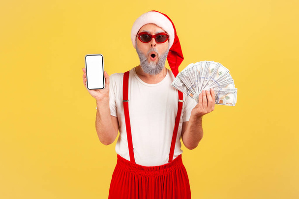 Excited shocked elderly man in santa claus hat and sunglasses holding dollar banknotes and empty screen smartphone, holiday bonuses, online banking. Indoor studio shot isolated on yellow background - Foto, afbeelding
