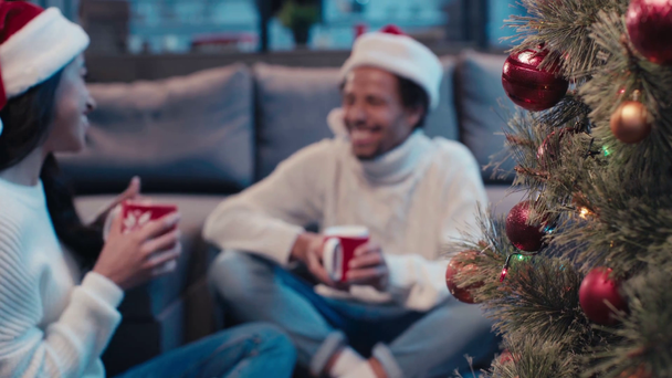 Christmas tree near african american couple with cups on blurred background - Footage, Video