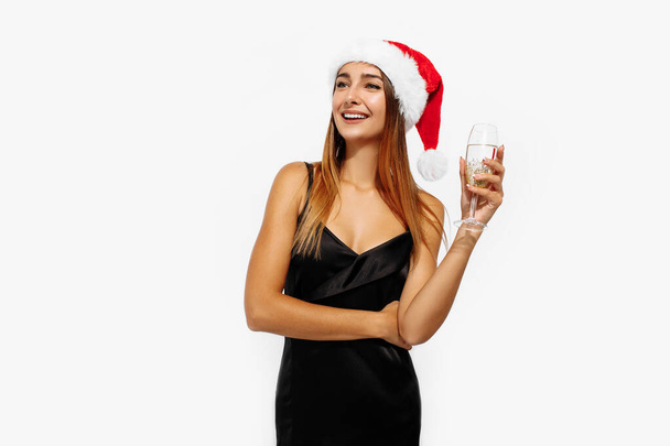Happy young woman, in a Christmas hat and an elegant black dress, holds a glass, on a white background, The concept of Christmas, holiday, New Year - Fotó, kép