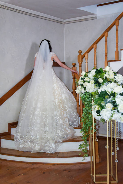 Hermosa novia posando en las escaleras de la habitación. Una novia en un vestido de novia blanco se encuentra en una escalera de madera. Novia en las escaleras de la casa. Una hermosa novia hermosa sube las escaleras. - Foto, imagen
