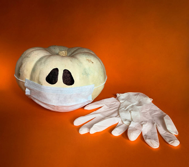 Nuovo concetto normale Halloween nella pandemia del COVID-19. Zucca indossando maschera protettiva e guanti medici accanto ad esso su uno sfondo arancione - Foto, immagini