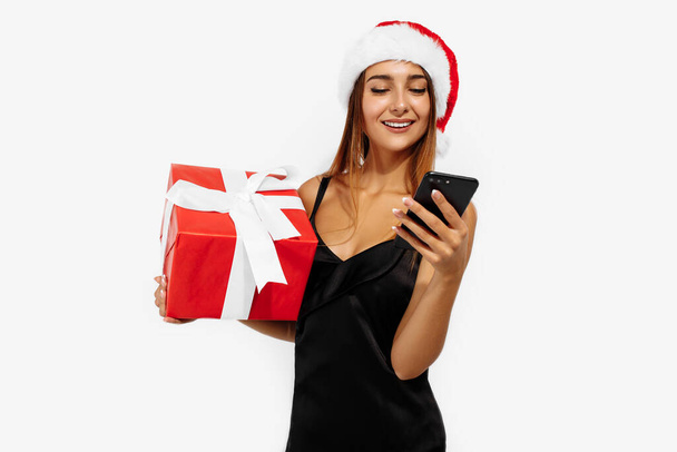 Mujer joven feliz en rojo sombrero de Navidad celebración caja de regalo y el uso de teléfono móvil aislado sobre fondo blanco - Foto, Imagen