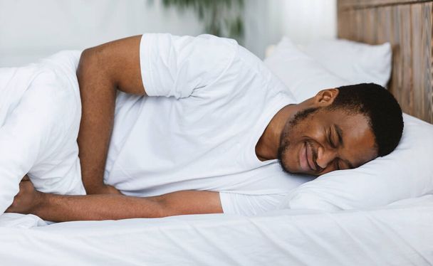 Αφρο-Αμερικανός άνθρωπος που έχει στομαχόπονο ξαπλωμένος στο κρεβάτι στο σπίτι - Φωτογραφία, εικόνα