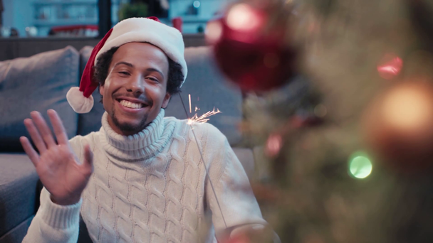 Noel Baba şapkalı mutlu Afro-Amerikan adam el sallıyor ve maytap tutuyor. - Video, Çekim