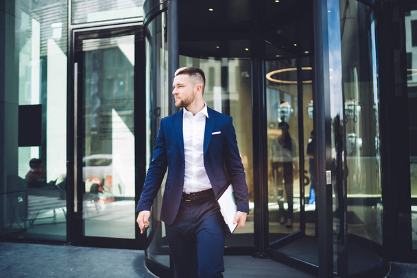 Elegant young male office worker in formal wear with clipboard leaving contemporary business centre building thoughtfully looking away in sunlight - Photo, Image