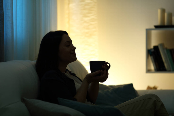 Profile silhouette of a woman drinking coffee at home in the dark night sitting on a couch - Fotoğraf, Görsel