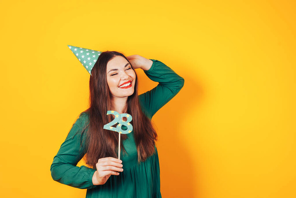 Красивая женщина празднует свой день рождения на желтом фоне в зеленом платье и на голове праздничный колпак держит номер 28 в руке. - Фото, изображение