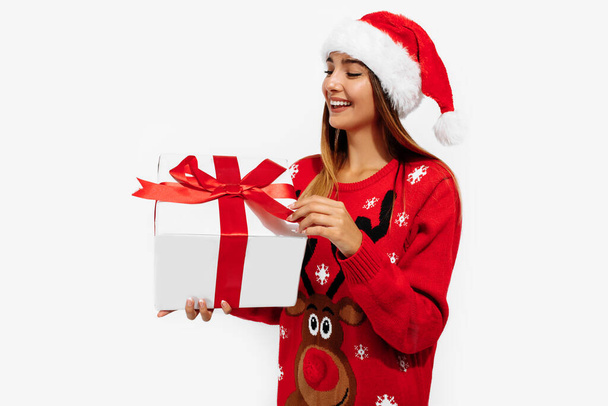 Cheerful young woman in a Christmas sweater and Santa Claus hat, with a gift on a white background, the concept of Christmas, New Year - Zdjęcie, obraz