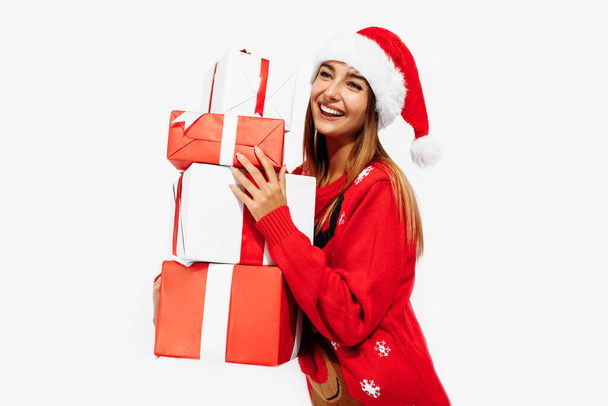 Happy brunette in red sweater and Santa Claus hat holding many gifts on white background, Christmas, holiday concept - Fotó, kép