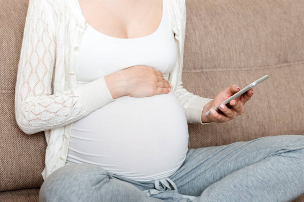 Καυκάσια έγκυος κοπέλα που χρησιμοποιεί κινητό τηλέφωνο, αναπαύεται στο κρεβάτι στο σπίτι. - Φωτογραφία, εικόνα