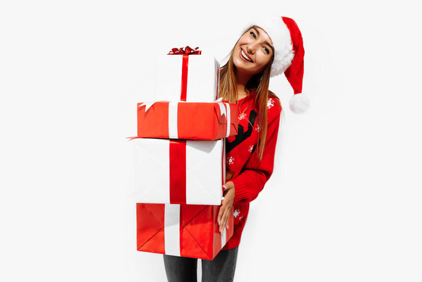 Happy beautiful brunette in red sweater and Santa Claus hat holding many gifts on white background, Christmas, holiday concept - Photo, Image