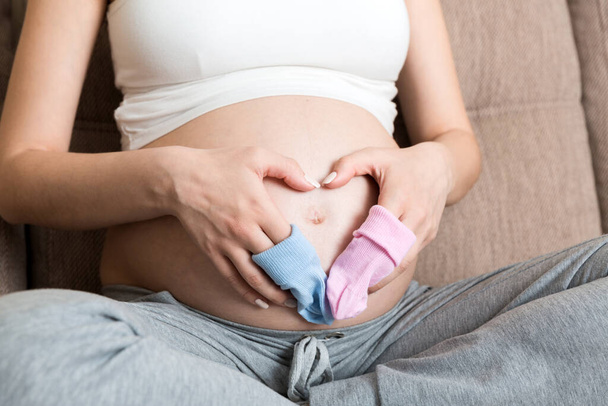 beautiful pregnant young woman holding a pair of cute baby socks on her tummy, lying on sofa at home. - Foto, imagen