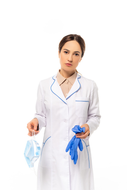 Doctor holding medical mask and latex gloves while looking at camera isolated on white - Foto, Bild