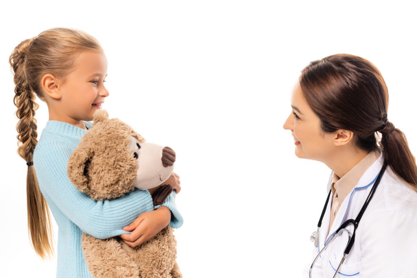 Smiling pediatrist looking at girl with soft toy isolated on white - Photo, Image