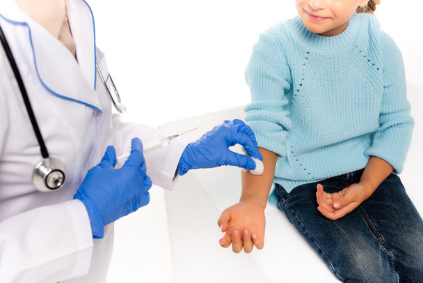 Ausgeschnittene Ansicht des Kinderarztes in Latexhandschuhen mit Baumwolle und Spritze in der Nähe der Hand des Kindes isoliert auf weiß - Foto, Bild