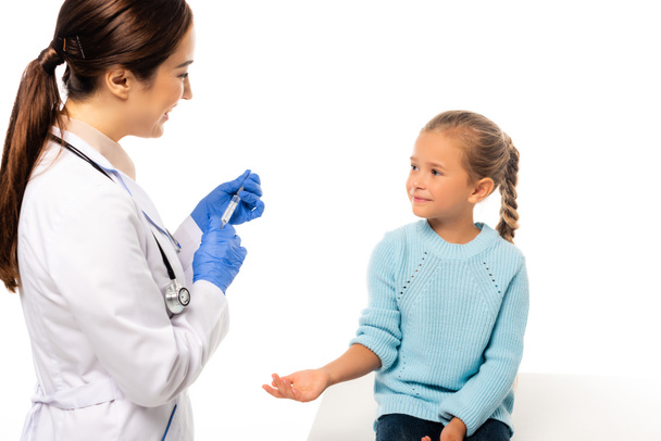 Smiling pediatrician in latex gloves holding syringe near child isolated on white - Zdjęcie, obraz