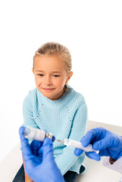 Selective focus of smiling girl looking at pediatrician holding syringe and vaccine isolated on white - Photo, image