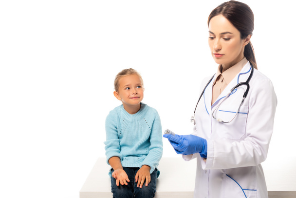 Врач в латексных перчатках держит вакцину рядом с испуганным ребенком изолированным на белом - Фото, изображение