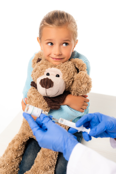 Concentration sélective d'une jeune fille souriante tenant un jouet souple près d'un pédiatre avec un vaccin et une seringue isolés sur du blanc - Photo, image