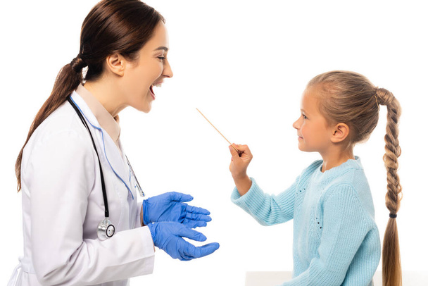 Smiling girl holding stick near pediatrician with open mouth isolated on white - Zdjęcie, obraz