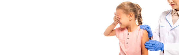 Панорамный снимок испуганного ребенка, закрывающего глаза рядом с доктором в латексных перчатках, изолированных на белом - Фото, изображение