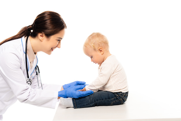Smiling pediatrician touching toddler boy on table isolated on white - Фото, изображение
