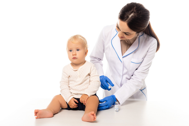Врач держит шприц с вакциной рядом с малышом на столе изолированы на белом - Фото, изображение