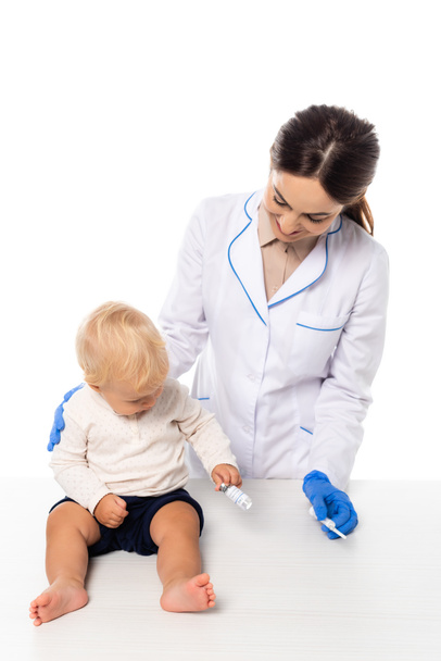 Un médecin souriant tenant une seringue près d'un tout-petit avec un pot de vaccin sur la table isolé sur blanc - Photo, image
