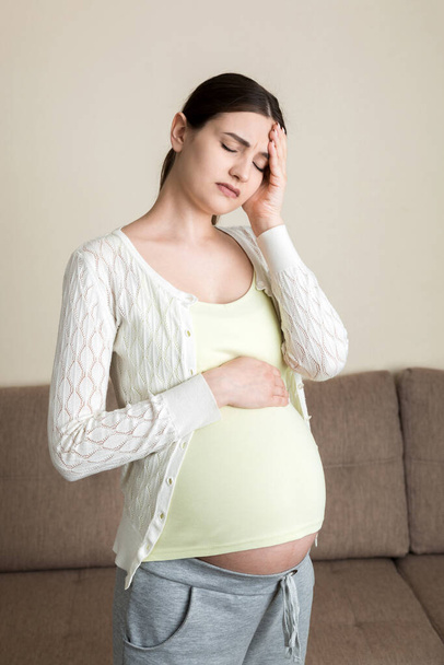Junge schwangere Frau mit Kopfschmerzen auf dem Sofa sitzend. Konzept Schwangerschaftserwartung. - Foto, Bild