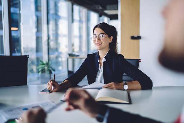 Portrait of happy female entrepreneur in optical eyewear for eyes protection smiling at camera during work cooperation with male colleague, successful business woman posing at desktop - 写真・画像
